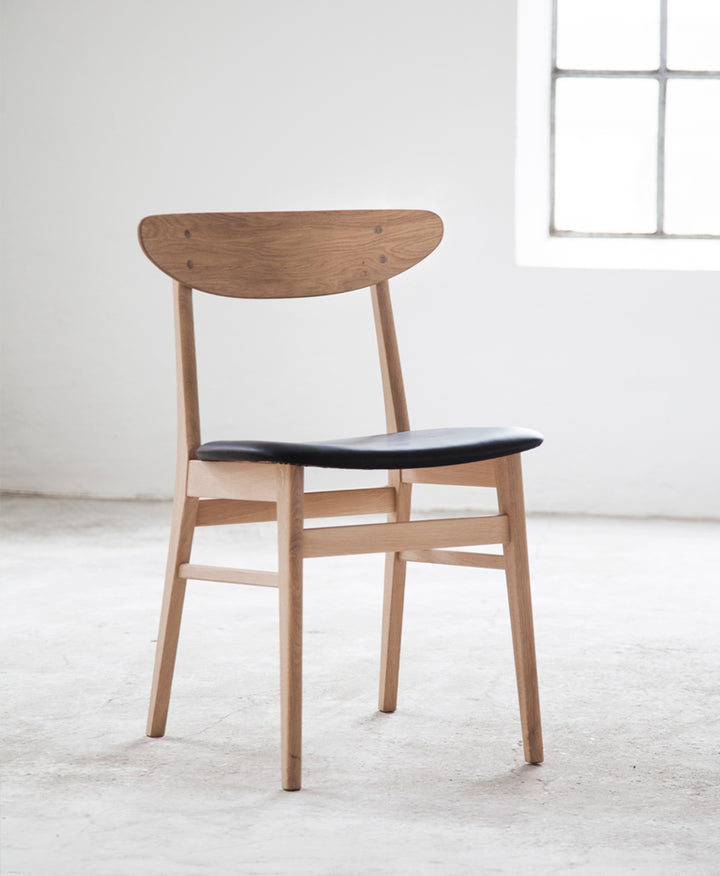 Klassisk spisebordsstol, model No. 210r, stel i natur eg og sædepolster i sort læder, stående i lyst miljø.
